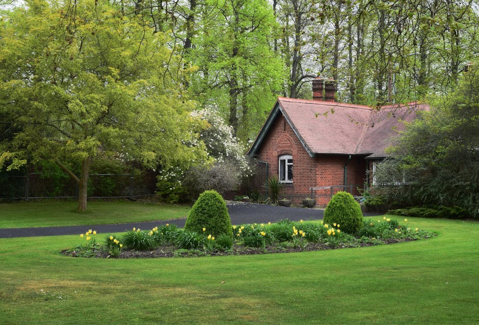 A well-maintained garden adjacent to a small red brick house featuring a pitched roof with red tiles and two brick chimneys. The foreground displays a lush green lawn with a circular flower bed border